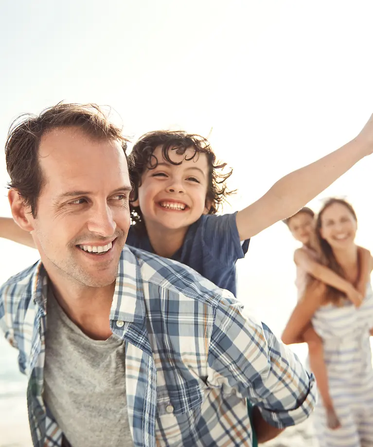 Mutter, Vater und deren zwei Kinder sind am Strand. Die Eltern tragen ihre Kinder Huckepack. 