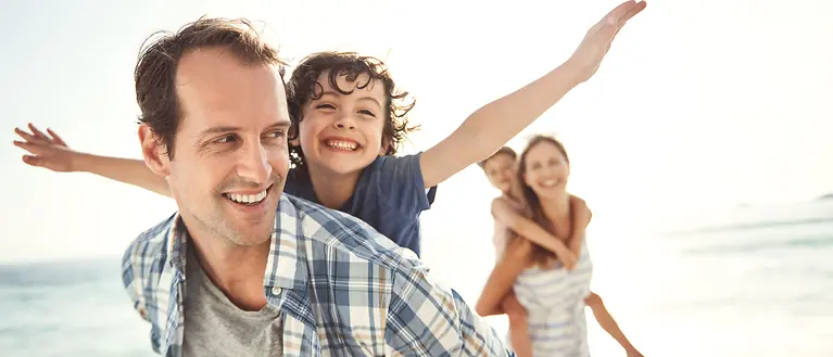 Mutter, Vater und deren zwei Kinder sind am Strand. Die Eltern tragen ihre Kinder Huckepack. 