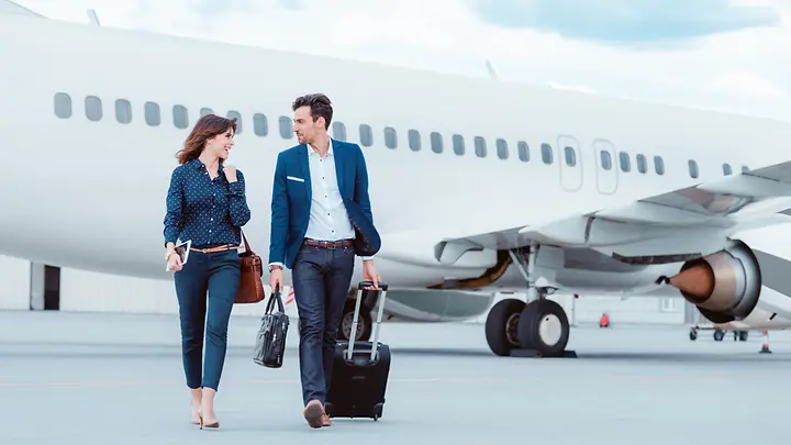 Eine Frau und ein Mann gehen auf dem Rollfeld am Flughafen. Im Hintergrund sieht man ein Flugzeug.