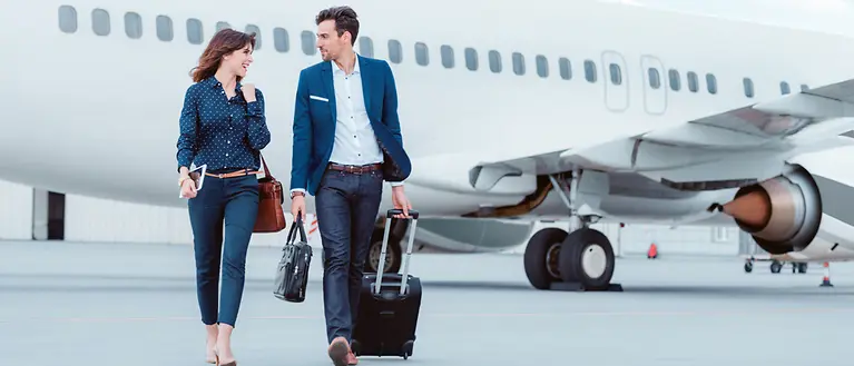 Eine Frau und ein Mann gehen auf dem Rollfeld am Flughafen. Im Hintergrund sieht man ein Flugzeug.