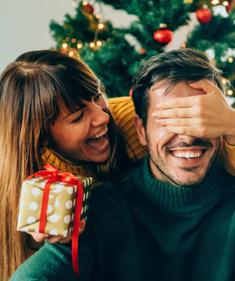 Eine Frau übergibt ihrem Mann ein Geschenk. Im Hintergrund ist ein geschmückter Weihnachtsbaum.