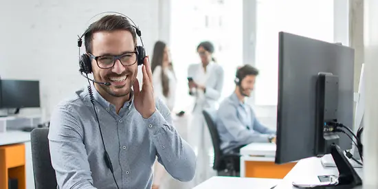 Ein Mann sitzt im Büro und trägt ein Headset. Im Hintergrund befinden sich noch weitere Mitarbeiter. 