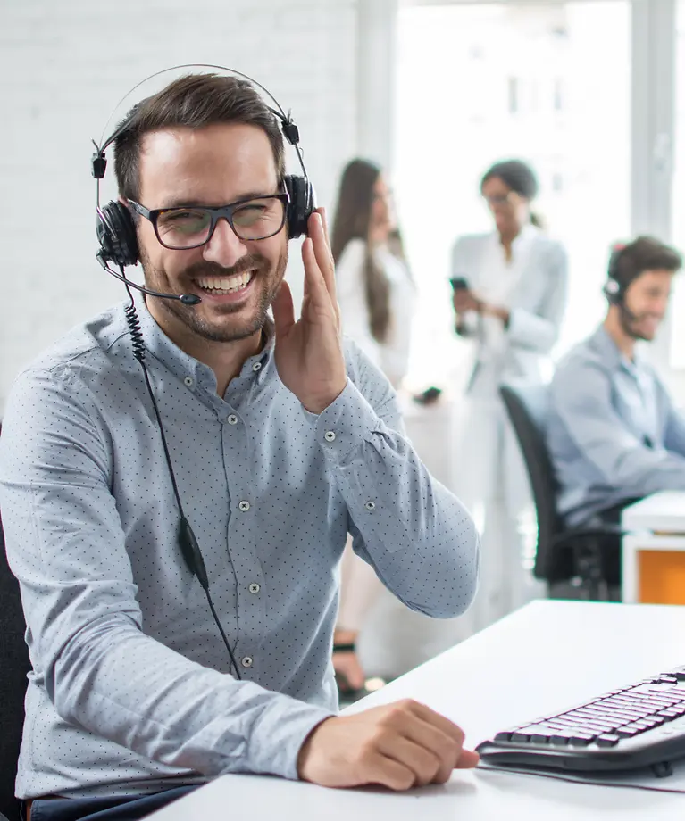 Ein Mann sitzt im Büro und trägt ein Headset. Im Hintergrund befinden sich noch weitere Mitarbeiter. 