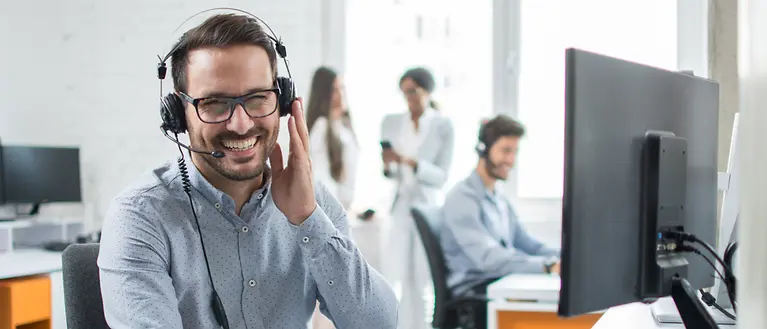 Ein Mann sitzt im Büro und trägt ein Headset. Im Hintergrund befinden sich noch weitere Mitarbeiter. 