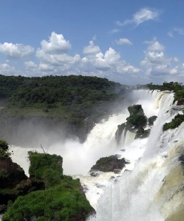 Man erkennt einen großen Wasserfall. Im Hintergrund sind viele Bäume und blauer Himmel zu sehen.