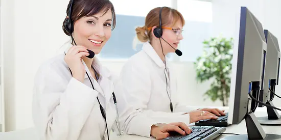 Zwei Frauen mit einem Headset sitzen am Computer und arbeiten. 