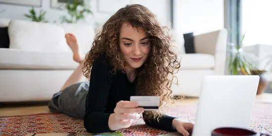 Eine Frau liegt auf dem Boden. Vor ihr steht ein Laptop. In ihrer Hand hat sie eine Kreditkarte. 