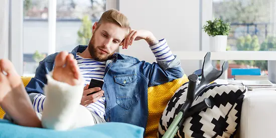 Ein Mann hat einen Gips am Fuß und sitzt auf dem Sofa. In seiner Hand hält er ein Handy.