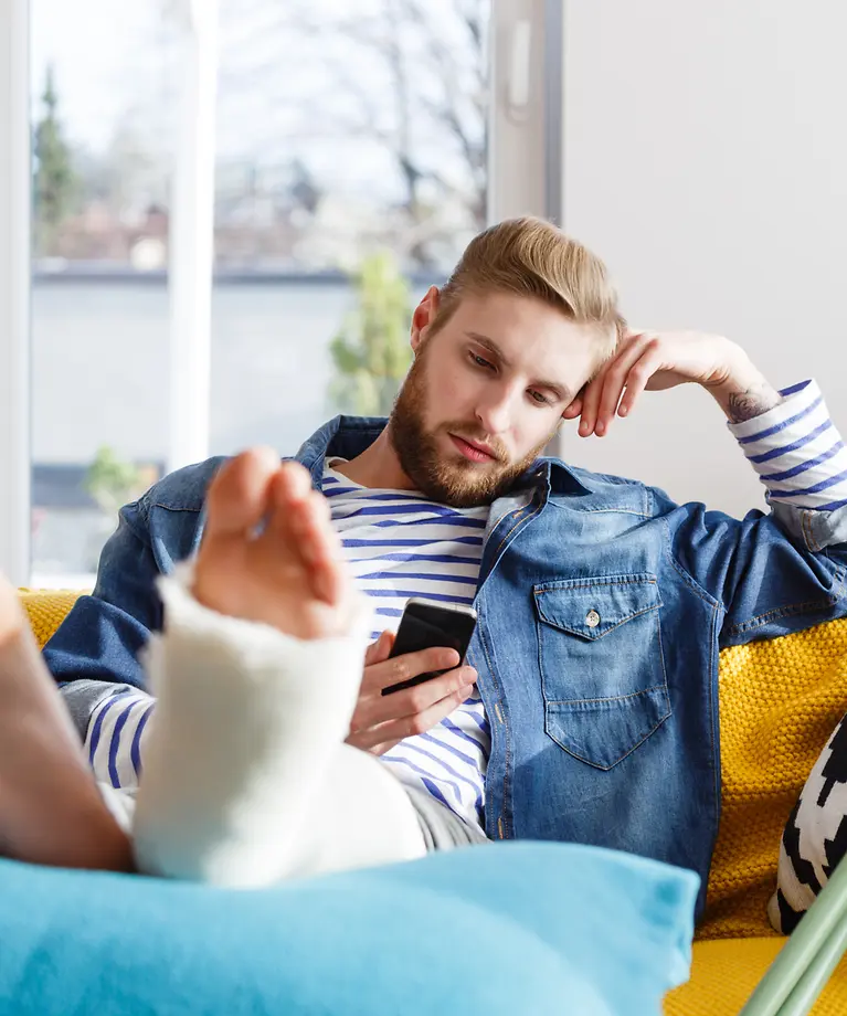 Ein Mann hat einen Gips am Fuß und sitzt auf dem Sofa. In seiner Hand hält er ein Handy.