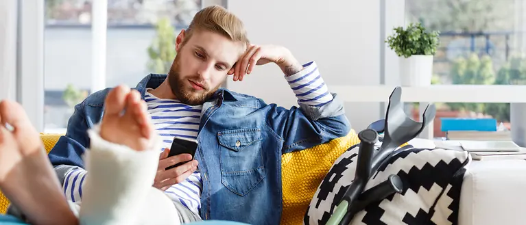 Ein Mann hat einen Gips am Fuß und sitzt auf dem Sofa. In seiner Hand hält er ein Handy.