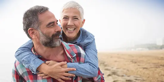 Eine Mann und eine Frau sind am Strand. Der Mann nimmt seine Frau Huckepack