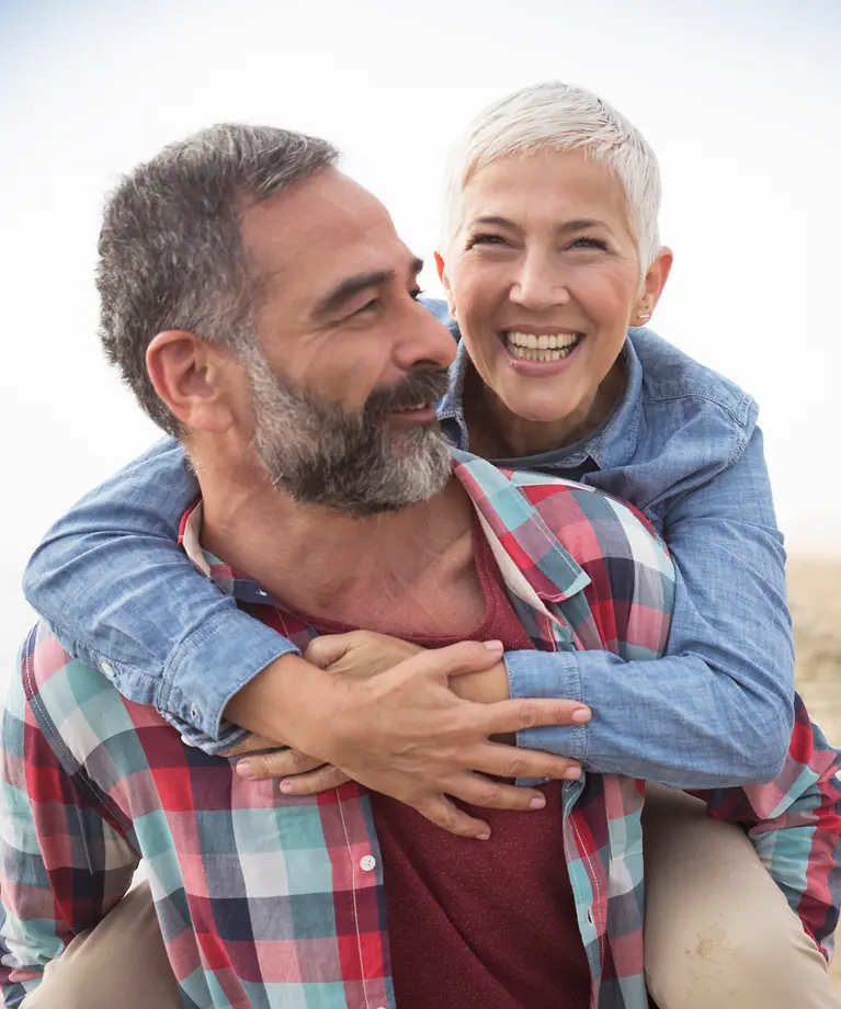 Eine Mann und eine Frau sind am Strand. Der Mann nimmt seine Frau Huckepack