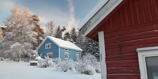 Man erkennt ein blaues Holzhaus umgeben von Schnee. Im Hintergrund sind schneebedeckte Bäume zu sehen.