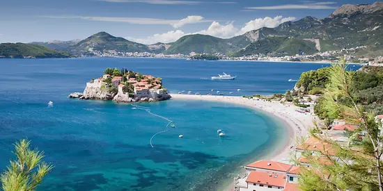 Man erkennt eine Bucht mit türkisem Wasser und Häusern direkt am Wasser. Im Hintergrund sind Berge zu sehen.