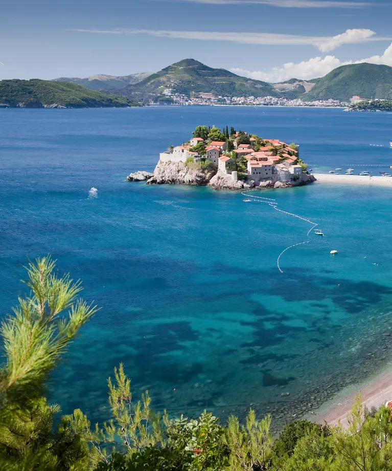 Man erkennt eine Bucht mit türkisem Wasser und Häusern direkt am Wasser. Im Hintergrund sind Berge zu sehen.