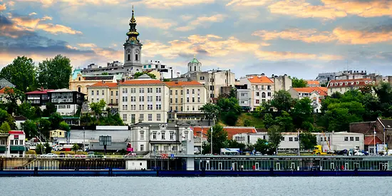 Man sieht eine kleine Stadt, die an einem Fluss liegt. Zwischen den Häusern wachsen grüne Bäume. 