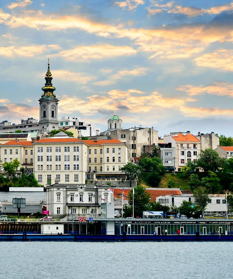 Man sieht eine kleine Stadt, die an einem Fluss liegt. Zwischen den Häusern wachsen grüne Bäume. 