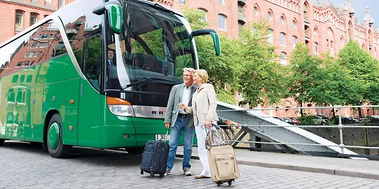 Zusehen sind zwei ältere Menschen, die mit ihren Koffern vor einem Bus stehen.