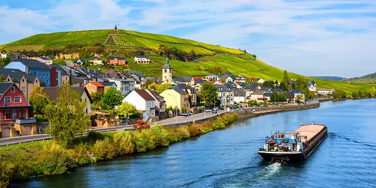 Man sieht ein Boot, dass auf der Mosel ist. Am Ufer stehen Häuser und es sind Weinberge zusehen.