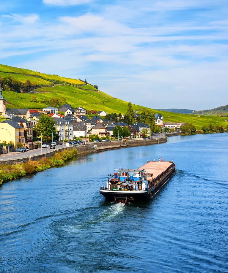 Man sieht ein Boot, dass auf der Mosel ist. Am Ufer stehen Häuser und es sind Weinberge zusehen.