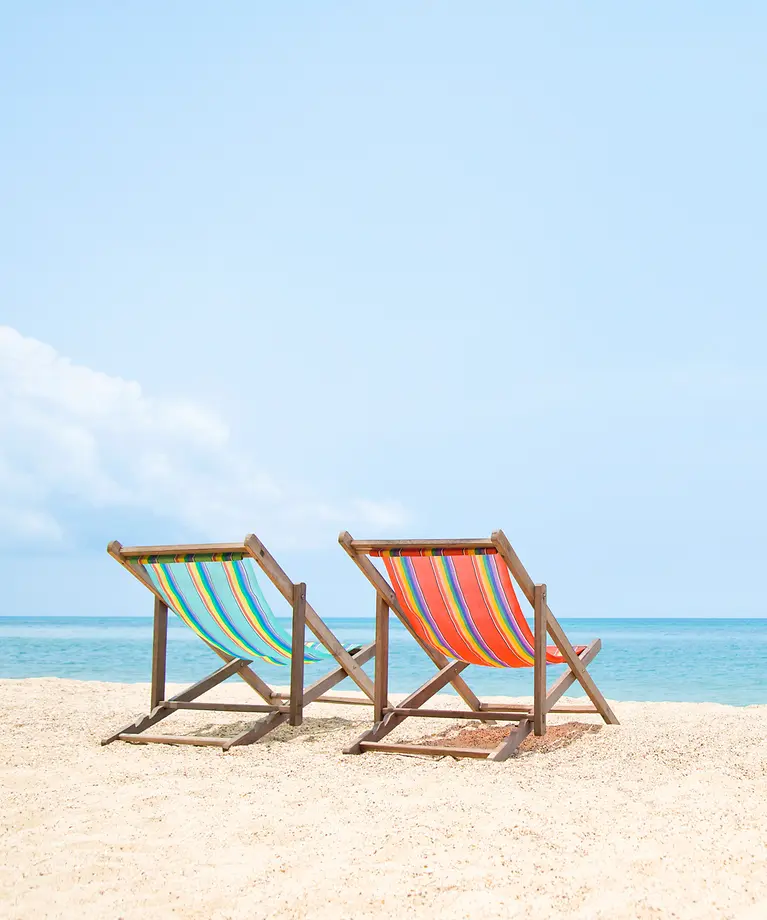 Zwei Liegestühle stehen am Strand. Im Hintergrund ist das Wasser.