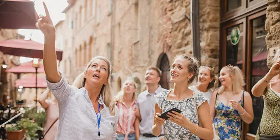 Eine Reisegruppe befindet sich in einer Altstadt. Eine Frau zeigt mit ihrem Finger auf ein Gebäude.