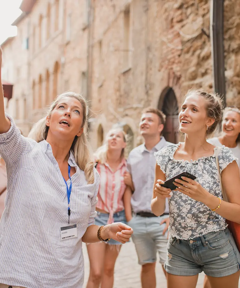 Eine Reisegruppe befindet sich in einer Altstadt. Eine Frau zeigt mit ihrem Finger auf ein Gebäude.