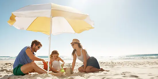 Eine Familie sitzt unter einem Sonnenschirm am Strand und spielen im Sand.