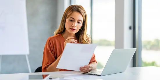 Eine Frau sitzt mit Papieren am Schreibtisch vor ihrem Laptop