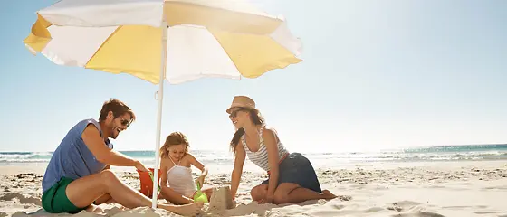 Familie am Strand - Reiseschutz Gold und Platin Eine Familie sitzt unter einem Sonnenschirm am Strand und spielen im Sand.