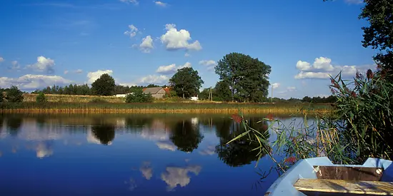 Man sieht ein See. Im Hintergrund sieht man grüne Bäume und ein Haus.