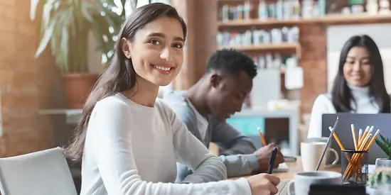 Drei Studenten sitzen an einem Tisch in einer Bibliothek und arbeiten. 