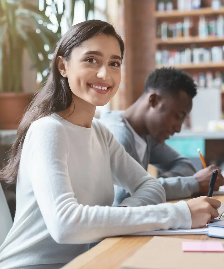 Drei Studenten sitzen an einem Tisch in einer Bibliothek und arbeiten. 