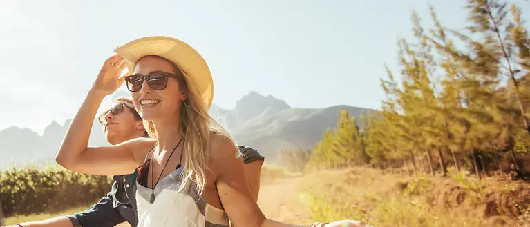 Eine Frau und ein Mann sitzen auf der Ladefläche eines Autos und genießen die Sonne. Im Hintergrund sind die Berge. 