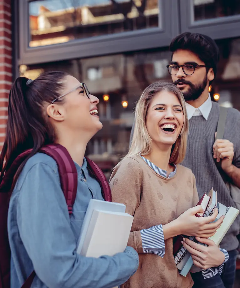 Drei Studenten mit Büchern in der Hand lachen. Im Hintergrund kann man ein Café sehen.