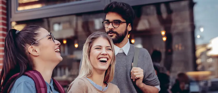 Drei Studenten mit Büchern in der Hand lachen. Im Hintergrund kann man ein Café sehen.